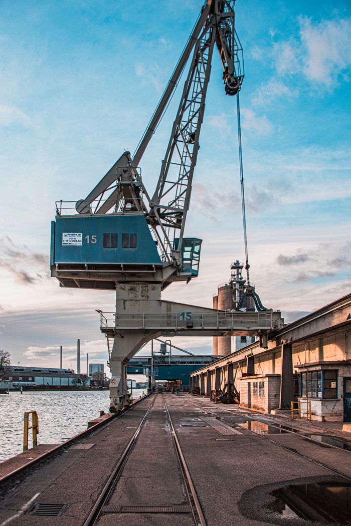 why-choose-us Vertical shot of a harbor crane by industrial warehouses during the day.