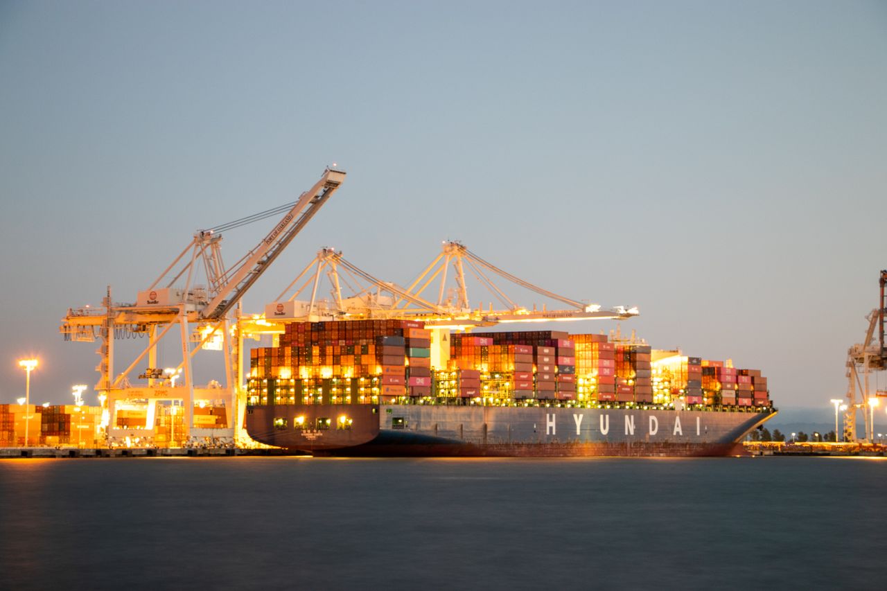 services-02 Container ship docked at port under cranes during twilight, illuminated against the evening sky.