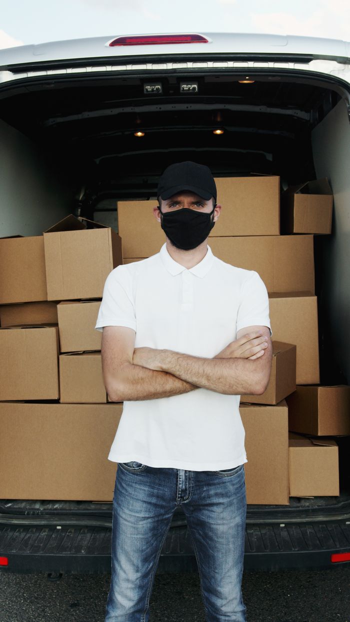 gallery-1 A courier wearing a mask standing in front of a loaded van with cardboard boxes, showcasing logistics.